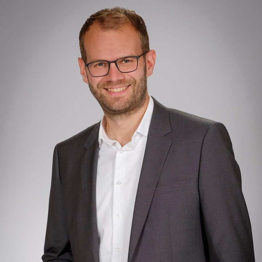 Mann im Anzug mit Brille lächelt freundlich vor neutralem Hintergrund.