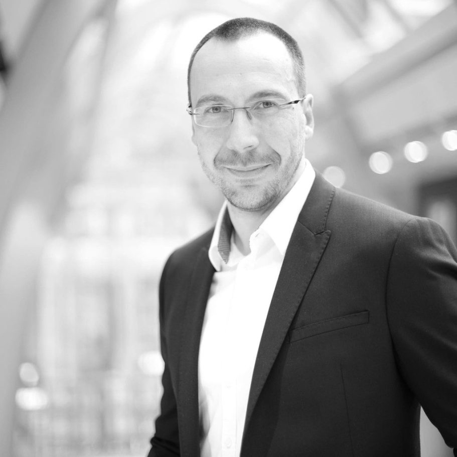 Lächelnder Mann in Anzug, mit Brille, vor verschwommenem Hintergrund.